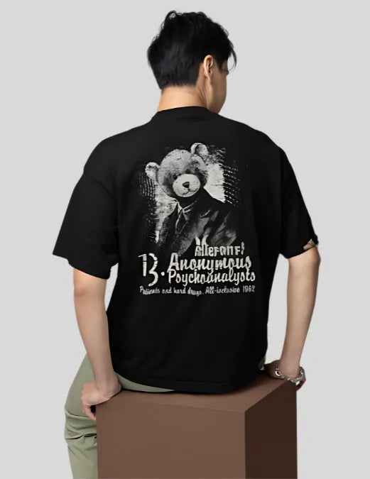 Person wearing a black t-shirt with a graphic design on the back, sitting on a box against a plain background.